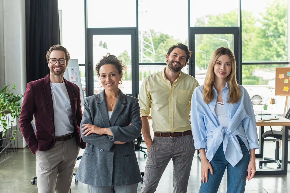 A diverse group of four young professionals stands confidently in a modern office with large windows. They are smiling, exuding a friendly, collaborative vibe.
