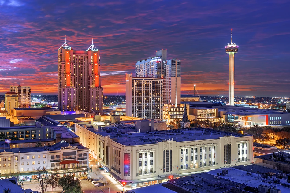 Cityscape of downtown San Antonio at dusk, featuring illuminated buildings and the Tower of the Americas under a vibrant purple and orange sky.