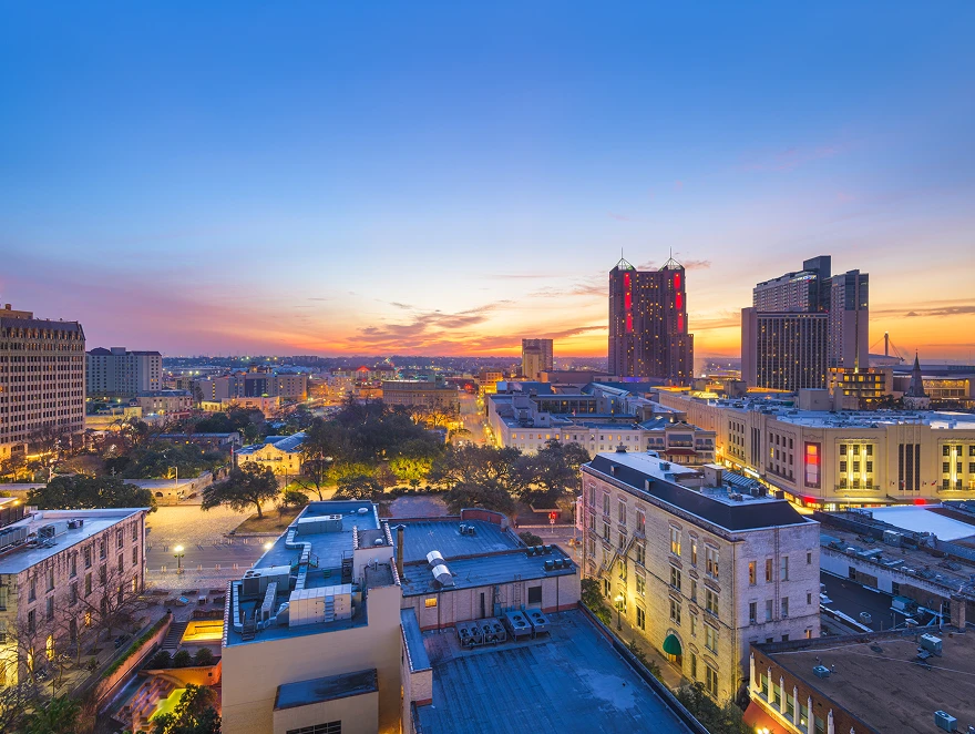 A vibrant cityscape at dusk with colorful skies, tall buildings with lights on, and tree-lined streets below. The sunset creates a warm, lively atmosphere.