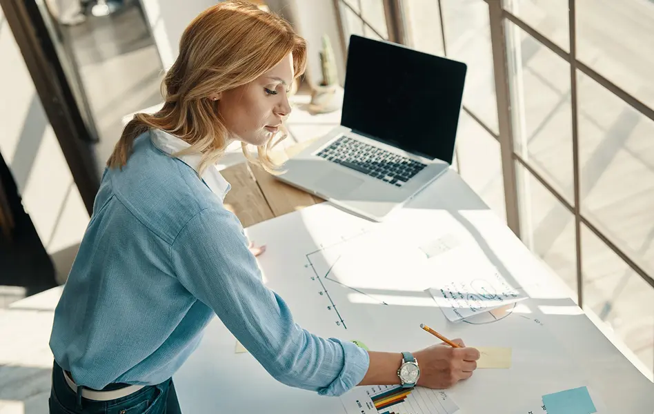 Woman working at a desk, reviewing documents and taking notes beside a laptop.