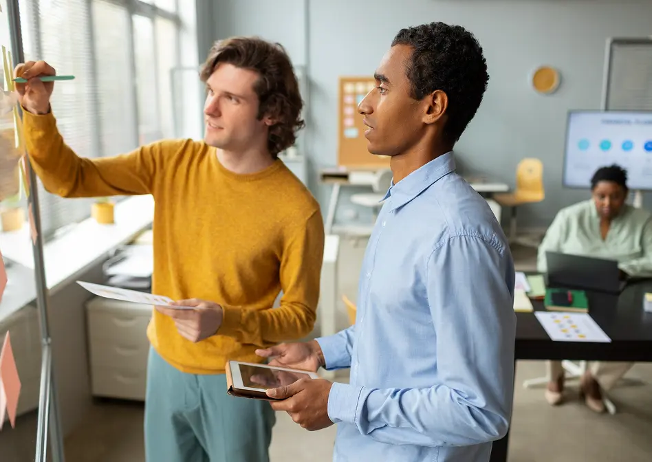 Two colleagues collaborating at a glass board with sticky notes in a modern office, one holding a tablet