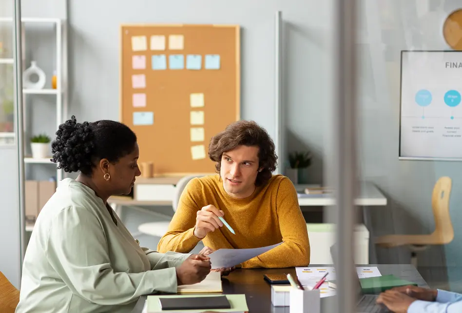 Two colleagues discussing documents at a desk in a modern office with a sticky note board and presentation screen in the background