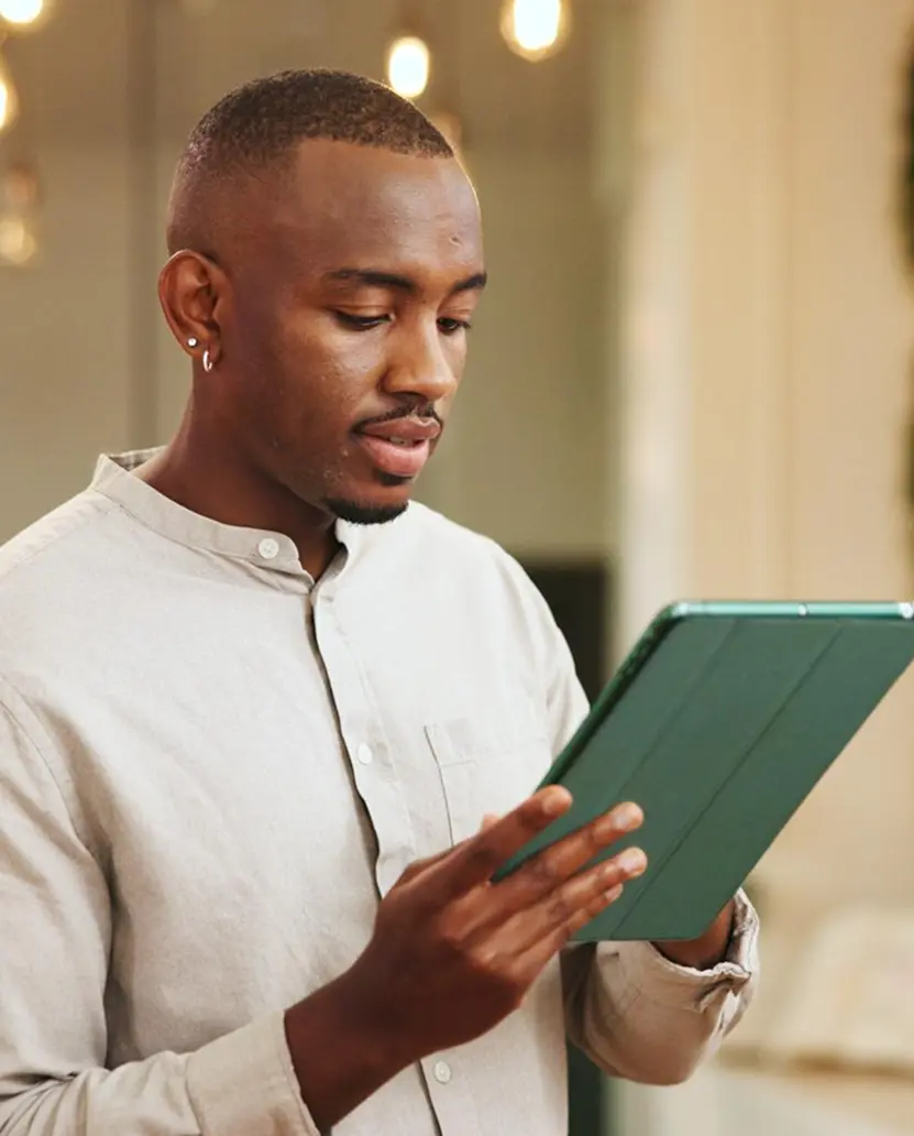 A young man in a beige shirt reading from a tablet with a green case in a softly lit interior