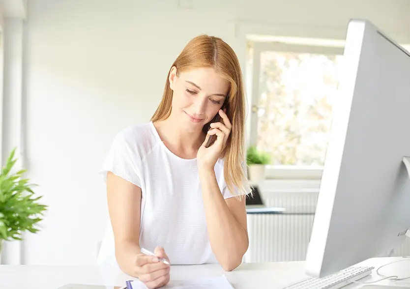 Woman talking on a phone while taking notes at a desk with a computer monitor